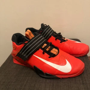 Nike savaleos Chile red powerlifting skor  - Säljer ett par riktigt snygga röda Nike power lifting skor med svart och vit randig rem över vristen. Skorna har svart snörning, vit swoosh och coola detaljer i orange och svart. Ovandelen är i syntetmaterial och mesh för extra komfort. Perfekta för dig som tränar och vill ha stil perfekta för gymmet för någon som tar det seriöst och ett riktigt bra pris då dessa slutat säljas och ny pris ligger på 2200 vid fler funderingar skriv till bara storlek 45 passar mer som 44