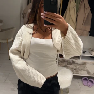 Vit croppad stickad kofta - Superfin vit croppad kofta med breda ärmar och ribbstickat material. Perfekt att slänga över ett linne för en chill och trendig look. Koftan har en öppen front och är riktigt mjuk och skön att bära.