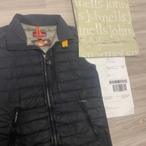 Svart dunväst från Parajumpers - Äkta Säljer en svart quiltad dunväst från Parajumpers med coolt gult detaljerat spänne vid kragen och två dragkedjeförsedda fickor. Västen har hög krage och är superlätt, perfekt för lager-på-lager. Insidan är grå och har snygga märkestaggar. Möts bara upp