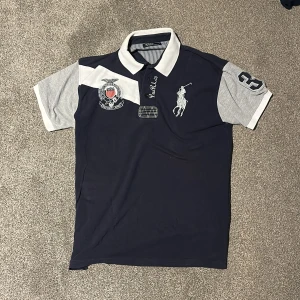 Piké från Ralph Lauren, marinblå - Snygg polo piké från Ralph Lauren i marinblått med gråa ärmar och vit krage. Stora broderade loggor och detaljer på bröstet och ärmen. Klassisk piké med knappar framtill, perfekt för en sportig och stilren look.