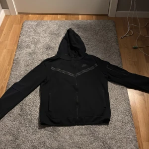 Svart Nike tech hoodie med dragkedja - Svart hoodie från Nike med hel dragkedja och huva. Snygg detalj med en mörkgrå linje över bröstet och diskret Nike-logga. Perfekt för chill dagar eller när du vill ha en sportig look. Långa ärmar och två fickor framtill.