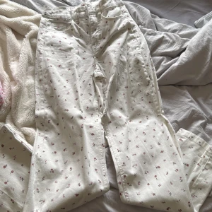 Blommiga vita jeansbyxor Denim - Säljer ett par vita jeansbyxor från Denim med sött blommigt mönster i rosa och rött. Byxorna har klassisk femficksdesign, dragkedja och knapp framtill samt bälteshällor. Perfekta för dig som vill ha en romantisk och trendig look.