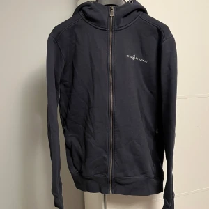 Svart hoodie från Sail Racing - Svart hoodie från Sail Racing med dragkedja framtill och broderad logga på bröstet. Tröjan har huva med tryckt detalj bak, långa ärmar och ribbade muddar. Perfekt för dig som gillar en clean och sportig stil.