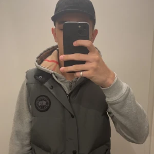 Black label canada goose väst - Säljer en mörkgrå dunväst från Canada Goose med hög krage och svart logotyp-patch på bröstet. Västen har knappar och dragkedja framtill samt två stora fickor. Perfekt för vintern! Storleken är M fast passar också S