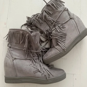 Grå mockaboots med fransar - Snygga grå boots i mocka med coola fransdetaljer och snörning framtill. Skorna har en platt sula och en diskret kilklack som ger extra höjd. Perfekta för dig som vill sticka ut med din stil.
