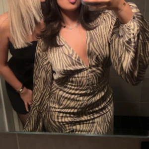 Zebramönstrad kortklänning från Adoore - Säljer en snygg kortklänning från Adoore med zebramönster i beige och brunt. Klänningen har v-ringning, ballongärmar och knappar framtill. Den är i storlek 40 men passar en 38 perfekt använd ca 3 gånger