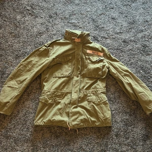 Grön parkajacka med fickor - Säljer en grön parkajacka i klassisk militärstil med fyra stora fickor framtill och hög krage med spänne. Jackan har dragsko i midjan och stängs med dragkedja. Perfekt för dig som gillar utility-vibe och streetstyle.