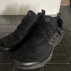 Svarta Nike Presto sneakers - Heltsvarta Nike Presto sneakers med mesh-ovandel och plastdetaljer på sidorna. Skorna har snörning, rund tå och platt sula för en sportig look. Perfekta för dig som gillar minimalistisk stil och vill ha bekväma sneakers till vardags.