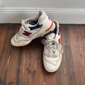 Vita sneakers från New Balance x Aimé Leon Dore - Snygga vita sneakers från New Balance i samarbete med Aimé Leon Dore. Skorna har röda och blå detaljer samt klassisk snörning. Perfekta för en stilren look med en touch av retro.