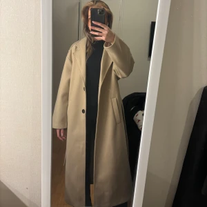 Beige kappa - Säljer en stilren beige kappa med knappar framtill. Den har en klassisk design med långa ärmar och en rak passform. Perfekt för att hålla sig varm och snygg under kyligare dagar.