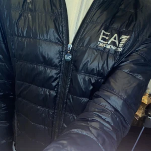 Svart dunjacka från Emporio Armani - Snygg svart dunjacka från Emporio Armani med EA7-logga på bröstet. Jackan har en dragkedja framtill och är perfekt för kyligare dagar. Den är lätt och stilren, vilket gör den enkel att matcha med olika outfits.passa på att skriva vid minsta fundering,priset går och diskutera. Stolek passar m passar S