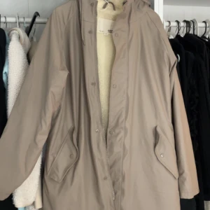 Beige regnjacka med fleecefoder - Säljer en beige regnjacka med mysigt vitt fleecefoder. Jackan har en huva och stängs med dragkedja och knappar. Perfekt för regniga dagar och håller dig varm och torr. 🧥