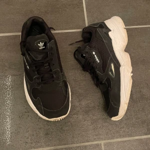 Svarta sneakers från Adidas - Säljer ett par svarta Adidas sneakers med vit sula. Skorna har snörning och den klassiska Adidas-loggan syns både på plösen och hälen. Skulle säga att dom är mellan 37/38 i storlek 