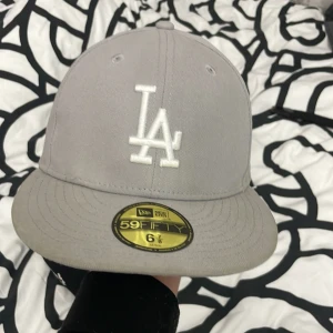 Grå keps från New Era - Snygg grå keps från New Era med broderad vit logga framtill. Modellen är 59FIFTY och har en klassisk passform. Perfekt för en stilren look.