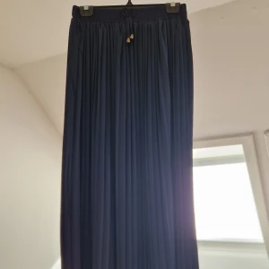 Mörkblå plisserad långkjol - Elegant mörkblå långkjol med plisserad design och elastisk midja. Perfekt för en stilren look. Kjolen har en diskret knytning i midjan för extra detalj.