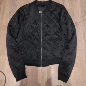 Svart quiltad bomberjacka - Säljer en stilren svart quiltad bomberjacka med dragkedja framtill. Jackan har långa ärmar och ribbade muddar vid ärmslut och nederkant. Perfekt för en cool och avslappnad look.