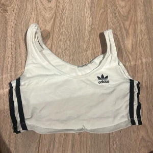 Vit linnetopp från Adidas - Säljer en vit linnetopp från Adidas med svarta ränder på sidorna och logga på bröstet. Perfekt för träning eller en avslappnad dag. Skön och enkel design.