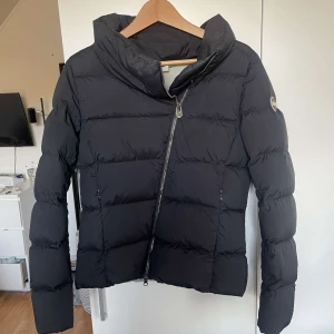 Svart dunjacka från Colmar - Snygg svart dunjacka från Colmar med dragkedja och hög krage. Perfekt till vår och höst då den är lite tunnare men funkar även på vintern. Jackan har en stilren design med Colmar-logga på ärmen. Den är i väldigt fint skick utifrån men lite fläckar inuti men som inte märks av när man har på sig den 