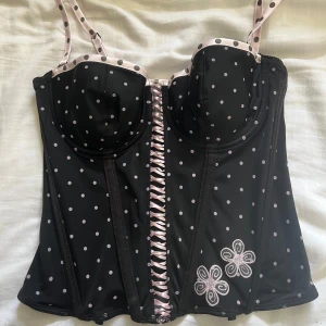 Svart prickig korsett  - Säljer en snygg svart korsett från Abecita med vita prickar och rosa detaljer. Den har justerbara axelband och snörning framtill. Perfekt för att ge en elegant touch till din outfit. Storlek 85b