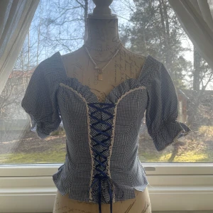 Blå och vit rutig blus med snörning - Charmig blå och vit rutig blus med snörning framtill och puffiga kortärmar. Perfekt för en somrig look med en söt och lekfull stil.