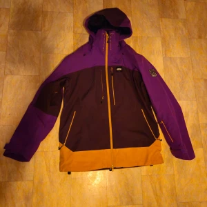 Lila och gul jacka från Picture - Säljer en lila och gul jacka från Picture. Jackan har en huva och flera praktiska fickor med dragkedjor. Perfekt för kyliga dagar med sin långa ärm och vindtäta design. Den har en unik färgkombination som verkligen sticker ut.