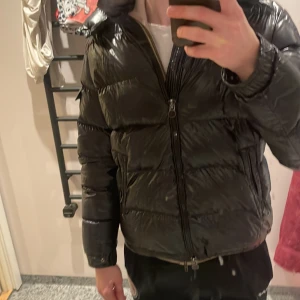 Svart dunjacka från Moncler - Säljer en snygg svart dunjacka från Moncler. Sliten inifrån men utifrån ser den bra ut som ni ser att spännet är trasig därav priset är billigare. Storlek 5 passar mig nästan lite för liten är typ 188 vid 75 kg. Hör av er för minsta lilla!