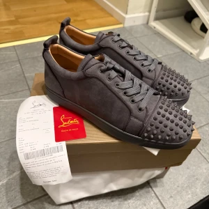 GRÅ LOUBS STORLEK 45 - Snygga grå sneakers från Christian Louboutin med nitar på tåpartiet. Skorna har en ikonisk röd sula och är tillverkade i mocka. Perfekta för att ge en edgy touch till din stil.