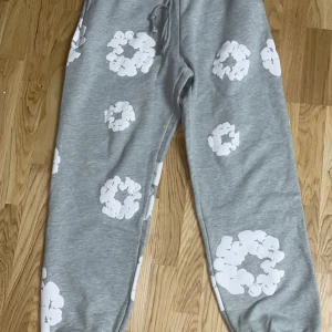 Grå mjukisbyxor från Denim Tears - Säljer ett par grå mjukisbyxor från Denim Tears med vita blommönster. Byxorna har en bekväm passform och dragsko i midjan för justerbarhet. Perfekta för en avslappnad stil.