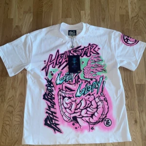 Vit t-shirt med grafiskt tryck från Hellstar - Cool vit t-shirt från Hellstar med ett färgstarkt grafiskt tryck i rosa, grönt och svart. Texten 'Hellstar' och 'No Guts No Glory' pryder både fram- och baksidan. Perfekt för dig som vill sticka ut med en unik stil.