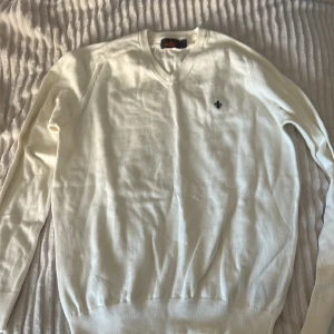 Vit tröja från Morris - Säljer en stilren vit tröja från Morris med v-ringning och ett diskret emblem på bröstet. Tröjan har långa ärmar och ribbade kanter vid ärmslut och nederkant. Perfekt för en klassisk look. Kan se skrinklyg ut på bild men den är hel ny