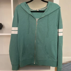 Grön hoodie från Victoria's Secret - Säljer en snygg grön hoodie från Victoria's Secret med dragkedja och huva. Den har vita ränder på ärmarna och texten 'ANGEL' på ryggen. Perfekt för en avslappnad stil. 💚