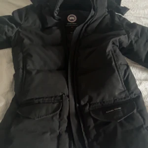 Svart dunjacka från Canada Goose - Säljer en svart dunjacka från Canada Goose. Jackan har en klassisk design med dragkedja och två stora fickor framtill. Perfekt för kalla vinterdagar med sin varma och isolerande fyllning. Ikonisk logga på ärmen.