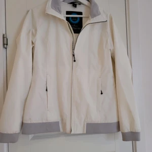 Vit jacka från XD Apparel - Snygg vit vindjacka från XD Apparel med dragkedja och gråa detaljer vid ärmslut och krage. Perfekt för en stilren look. Jackan har en praktisk hörlursöppning på insidan.