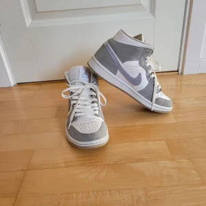 Grå, blå och vita Nike sneakers - Snygga grå, blå och vita Nike Jordan 1 sneakers med klassisk design. Skorna har snörning och en stilren färgkombination med blå Nike-logga på sidan. Perfekta för en avslappnad stil.
