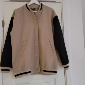 Beige och svart bomberjacka med text - Snygg beige bomberjacka med svarta ärmar och texten 'AUTHENTIC' på ryggen. Jackan har knappar framtill och ribbade muddar i svart och beige. Perfekt för en cool och avslappnad stil.