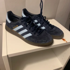 Adidas Handball Spezial i mörkblå mocka - Snygga Adidas Handball Spezial sneakers i mörkblå mocka med ljusblå ränder. Skorna har en klassisk design med gummisula och snörning. Perfekta för en sportig look.