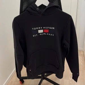 Svart hoodie från Tommy Hilfiger - Säljer en stilren svart hoodie från Tommy Hilfiger med logotyp framtill och på huvan. Tillverkad i ekologisk bomull, perfekt för en avslappnad stil. Hoodien har en klassisk känguruficka och ribbade muddar vid ärmslut och nederkant.