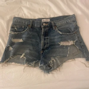 Blå jeansshorts från Zara - Säljer ett par coola blå jeansshorts från Zara med slitna detaljer och fransar. De har en klassisk femficksdesign och knappgylf. Perfekta för en avslappnad stil.