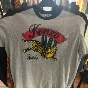 Grå t-shirt från Kenzo - Säljer en grå t-shirt från Kenzo som inköptes på NK 2017. Sparsamt använd då jag har växt ur den. Trycket är deras gamla logga och detta är för något jubileum.