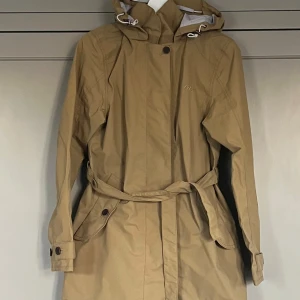 Didriksons Beige jacka.  - Snygg beige jacka med huva och knappar. Jackan har ett bälte i midjan och långa ärmar med knappar vid ärmsluten. Perfekt för vår och höst! Stormsystem- waterproof. 
