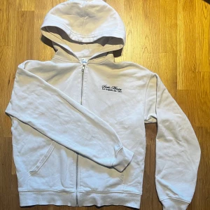 Santa Monica hoodie från H&M - Säljer en vit hoodie från H&M med texten 'Santa Monica, Los Angeles, Est. 1991' broderad på bröstet. Den har dragkedja och en bekväm huva. Perfekt för en avslappnad stil.