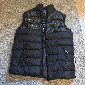 Svart dunväst från Philipp Plein - Snygg svart dunväst från Philipp Plein med broderad logga på bröstet. Västen har en dragkedja framtill och är perfekt för kyligare dagar. Tillverkad i Italien med hög kvalitet och stilren design.