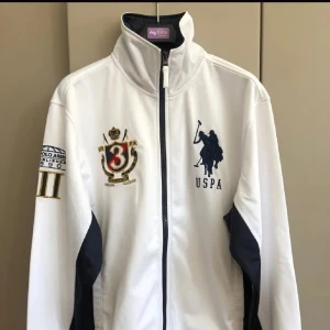 Vit jacka/zip tröja  från USPA - Säljer en stilren vit jacka från USPA med dragkedja och broderade detaljer på bröstet. Jackan har långa ärmar och mörkblå inslag på sidorna och insidan av kragen. Perfekt för en sportig look!