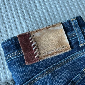 Blå jeans från Jacob Cohën - Snygga blå jeans från Jacob Cohën med unika detaljer som en läderpatch med snörning på baksidan. Jeansen har en klassisk femficksdesign och knappar framtill. Perfekta för en stilren look.