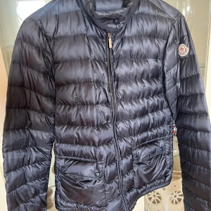 Svart dunjacka från Moncler - Snygg svart dunjacka från Moncler med dragkedja och två fickor framtill. Jackan har en quiltad design och en hög krage för extra värme. Perfekt för kyliga dagar. Finns tecken på användning - ett igensytt hål på rygg + inuti jackan som knappt syns. Nyss kemtvättad & lagat dragkedjan hos skräddare. Storlek 1