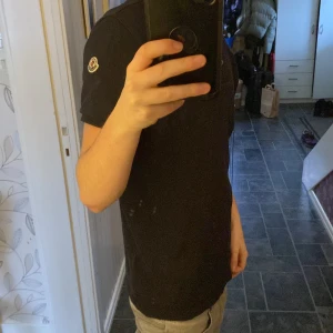 Svart kortärmad pikétröja från Moncler - Säljer en stilren svart pikétröja från Moncler. Tröjan har en klassisk krage med knappar och korta ärmar. Perfekt för en avslappnad men ändå snygg look. Säljer billigt för den har ett hål