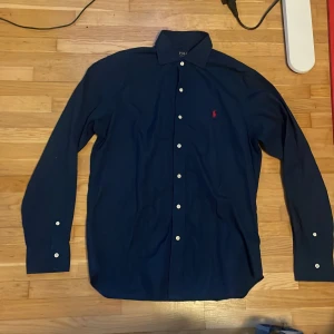  Polo Ralph Lauren skjorta  - Snygg mörkblå skjorta från Polo Ralph Lauren med klassisk krage och vita knappar. Den har en broderad logga i rött på bröstet och långa ärmar. Perfekt för en stilren look.