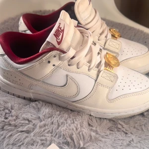 Vita och röda Nike sneakers - Snygga vita Nike sneakers med röda detaljer och gyllene dekorationer på snörningen. Skorna har en klassisk design med perforerade detaljer på tån och en bekväm passform. Perfekta för en stilren look.