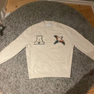 Vit sweatshirt från Axel Arigato - Säljer en stilren vit sweatshirt från Axel Arigato med coola applikationer på bröstet. Tröjan har långa ärmar och en rund halsringning. Perfekt för en avslappnad look. Nypris 2300kr
