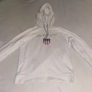 Vit hoodie från Gant - Säljer en stilren vit hoodie från Gant med ett tryck av märkesnamnet och en sköld med stjärnor och ränder på bröstet. Hoodien har en justerbar huva med snören och långa ärmar. Perfekt för en avslappnad stil.